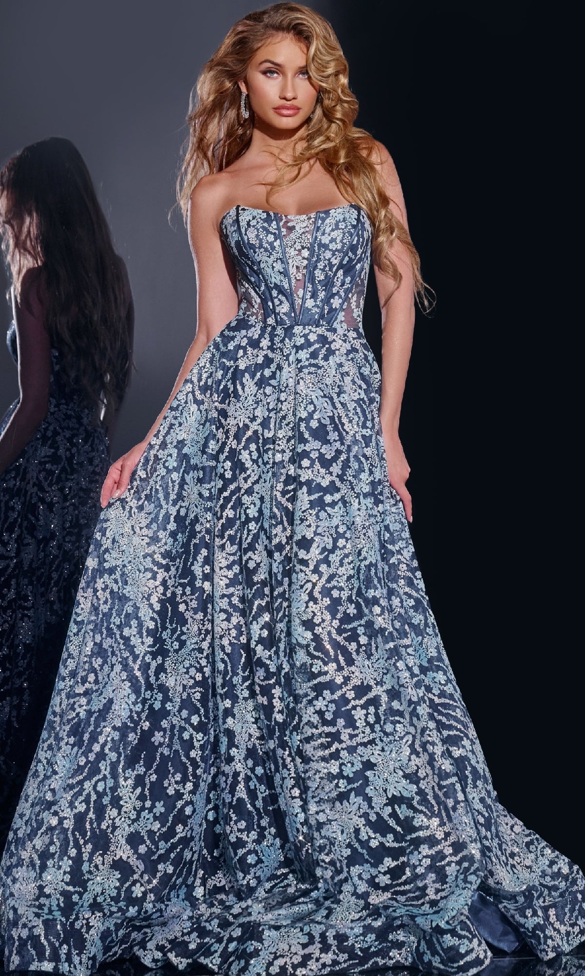 Wearing the Jovani 44151 long prom dress, a woman stands against a dark background. The strapless navy gown features a corset bodice and intricate silver embroidery, with her reflection visible.